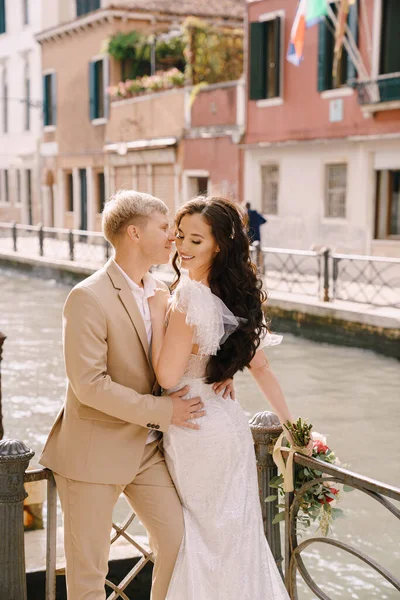 Italië bruiloft in Venetië. Pasgetrouwden staan aan de oevers van het kanaal van Venetië. De bruidegom knuffelt de bruid bij de taille. Witte trouwjurk met kleine mooie trein en zandkleurige heren pak. — Stockfoto
