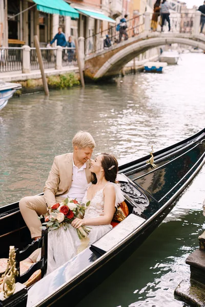 Italy wedding in Venice. The bride and groom ride in a classic wooden gondola along a narrow Venetian canal. Close-up of cuddles newlyweds. — Stock Photo, Image