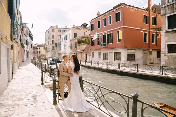 Італійське весілля у Венеції. Молоді люди стоять склавши руки на березі Венеційського каналу. Наречений обіймає наречену за талію. Біле весільне плаття з красивим поїздом і піщаний костюм.. — стокове фото