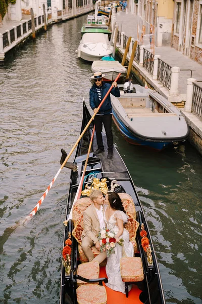 Italia boda en Venecia. Un gondolero enrolla a los novios en una clásica góndola de madera a lo largo de un estrecho canal veneciano. Los recién casados están sentados en un barco en el fondo de edificios antiguos . — Foto de Stock