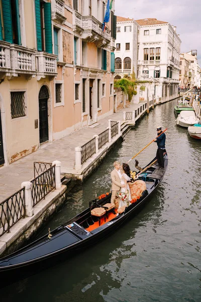 Italia boda en Venecia. Un gondolero enrolla a los novios en una clásica góndola de madera a lo largo de un estrecho canal veneciano. Los recién casados están de pie en un barco en el fondo de edificios antiguos . — Foto de Stock