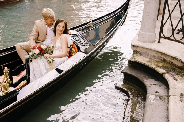 Olaszország esküvője Velencében. A menyasszony és a vőlegény lovagolni egy klasszikus fa gondola mentén egy keskeny velencei csatorna. Az újdonsült házasok ölelésének közelsége. — Stock Fotó