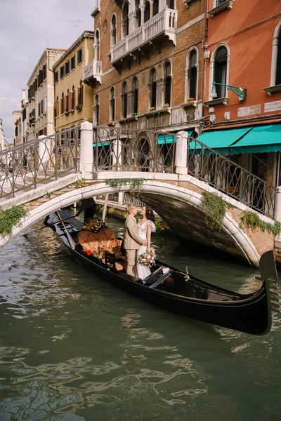 Італійське весілля у Венеції. Гондольєр кидає наречену з нареченим у класичну дерев'яну гондолу по вузькому венеціанському каналу. Молоді люди в гондолі пливуть з - під моста, стоять у човні і обіймають. — стокове фото