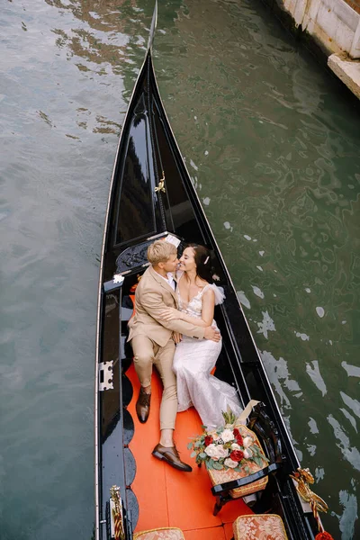 Ιταλικός γάμος στη Βενετία. Ένας γονδολιέρης τυλίγει μια νύφη και τον γαμπρό σε μια κλασική ξύλινη γόνδολα κατά μήκος ενός στενού βενετσιάνικου καναλιού. Οι νεόνυμφοι κάθονται σε μια βάρκα στο βάθος αρχαίων κτιρίων.. — Φωτογραφία Αρχείου