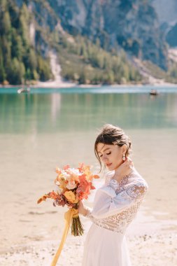Kolları ve dantelli beyaz elbiseli güzel bir gelin, İtalya 'da Lago di Braies' de sarı bir sonbahar buketi ve şakayık gülleri ile. Düğün Avrupa 'da, Braies Gölü' nde..
