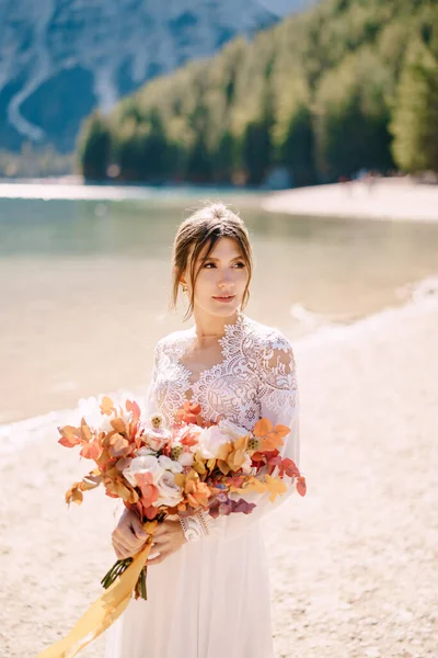 이탈리아 의 라고 디 브라이아 에는 소매와 레이스 가 달린 하얀 드레스를 입은 아름다운 신부가 마른 꽃 과 조랑말 장미로 누렇게 가을 부케 장식을 하고 있다. 유럽의 브레 이브스 호수에서의 목적지 결혼식. — 스톡 사진