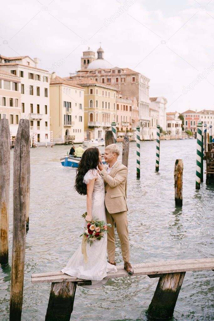 Italy wedding in Venice. The bride and groom are standing on a wooden pier for boats and gondolas, near the Striped green and white mooring poles, against backdrop of facades of Grand Canal buildings.