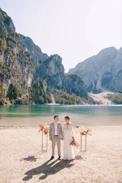 A noiva e o noivo estão no lugar para a cerimônia, com um arco de colunas de flores do outono, contra o pano de fundo do Lago di Braies, na Itália. Casamento de destino na Europa, no lago Braies . — Fotografia de Stock