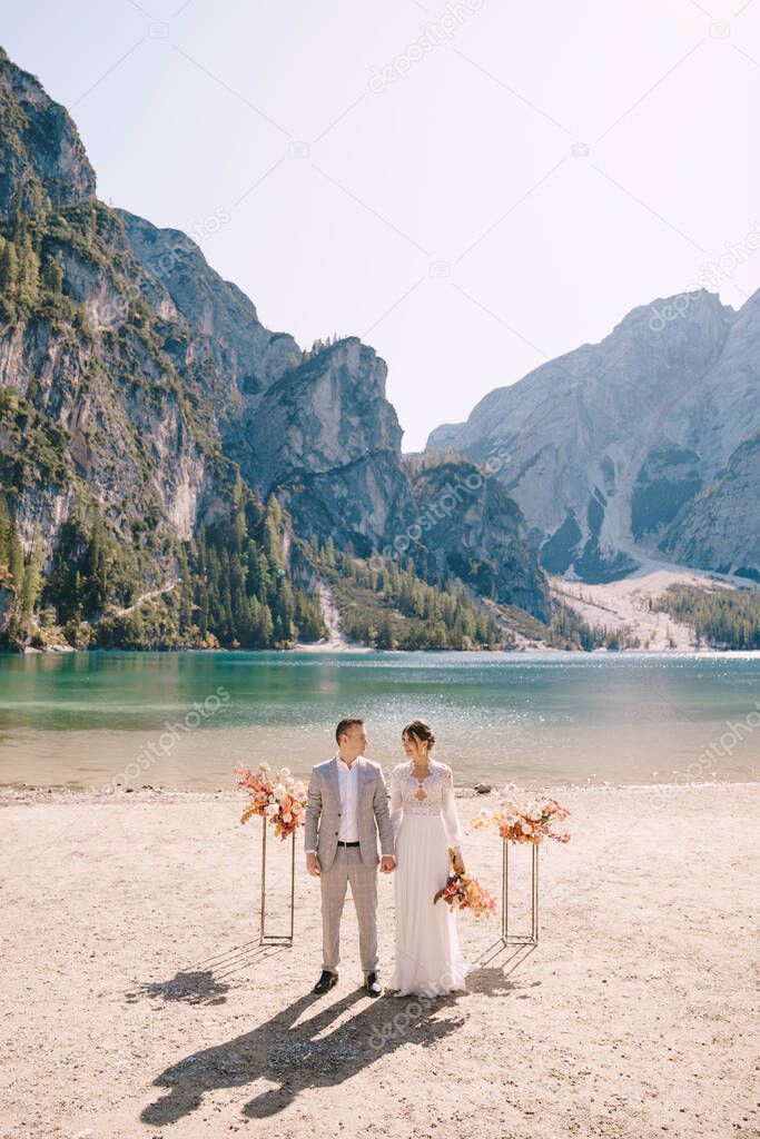 The bride and groom stand in place for the ceremony, with an arch of autumn flower columns, against the backdrop of the Lago di Braies in Italy. Destination wedding in Europe, on Braies lake.