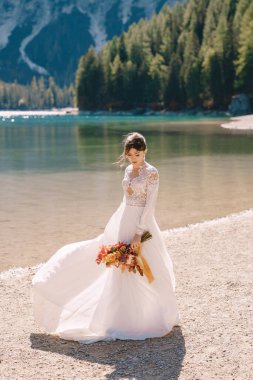 Kolları ve dantelli beyaz elbiseli güzel bir gelin, İtalya 'da Lago di Braies' de sarı bir sonbahar buketi ve şakayık gülleri ile. Düğün Avrupa 'da, Braies Gölü' nde..