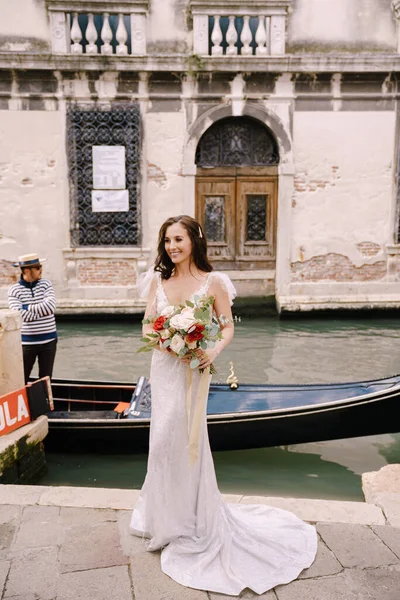 Velence, Olaszország - 2019. október 04.: Velencei esküvő Olaszországban. Egy fehér ruhás menyasszony, egy vonattal, csokorral a kezében, a mólón áll a kikötött gondola mellett egy keskeny velencei csatornában.. — Stock Fotó