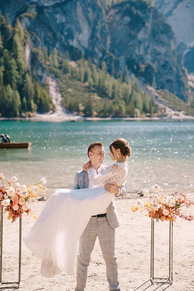 O noivo circunda a noiva em seus braços no local para a cerimônia, com duas colunas de flores de outono em vez de um arco, contra o pano de fundo do Lago di Braies, na Itália. Casamento na Europa, no lago Braies . — Fotografia de Stock