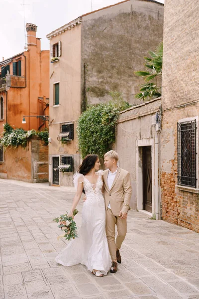 Mariage italien à Venise. Les mariés marchent le long des rues désertes de la ville. Les jeunes mariés embrassent, dansent, se tiennent la main sur la toile de fond de maisons pittoresques en briques rouges . — Photo