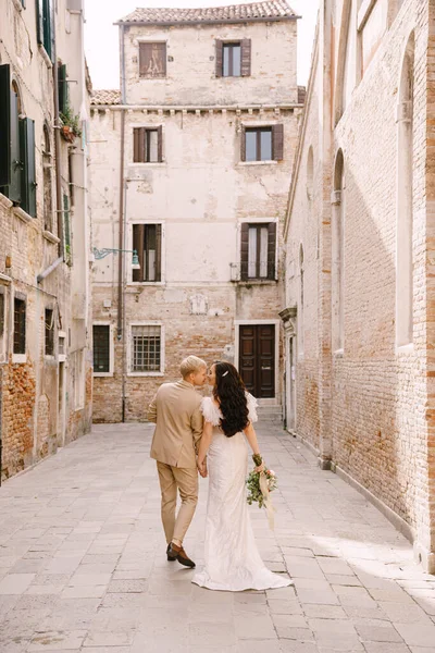 Olaszország esküvője Velencében. A menyasszony és a vőlegény a város elhagyatott utcáin sétálnak. A friss házasok egy zsákutcában sétálnak a téglaépületek hátterében.. — Stock Fotó