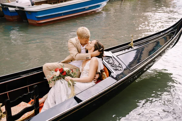 Olaszország esküvője Velencében. A menyasszony és a vőlegény lovagolni egy klasszikus fa gondola mentén egy keskeny velencei csatorna. Az újdonsült házasok ölelésének közelsége. — Stock Fotó