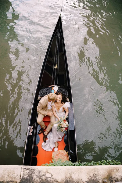 Olaszország esküvője Velencében. A gondolás gurít egy menyasszony és a vőlegény egy klasszikus fa gondola mentén egy keskeny velencei csatorna. A friss házasok egy csónakban ülnek az ősi épületek hátterében.. — Stock Fotó