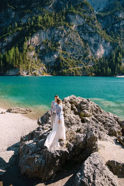 Les mariés se tiennent debout sur des pierres surplombant le lac de Braies en Italie. Mariage de destination en Europe, sur le lac de Braies. Aimer les jeunes mariés marcher dans le contexte de la nature étonnante . — Photo