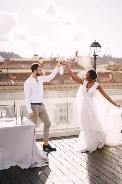 Διαφυλετικό ζευγάρι γάμων. Destination fine-art γάμος στη Φλωρεντία, Ιταλία. Αφροαμερικανή νύφη και Καυκάσιος γαμπρός χορεύουν κοντά στο τραπέζι για ένα γαμήλιο δείπνο, στην οροφή του εστιατορίου. — Φωτογραφία Αρχείου