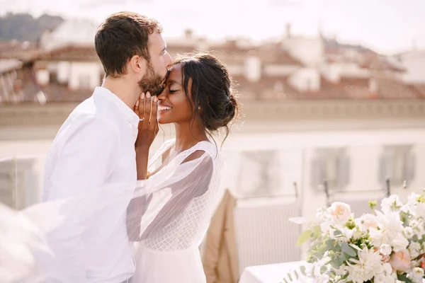 Tujuan pernikahan seni rupa di Florence, Italia. Pengantin pria kaukasia dan pengantin wanita Afrika-Amerika berpelukan di atap saat matahari terbenam. Pasangan pernikahan multiras Stok Gambar Bebas Royalti