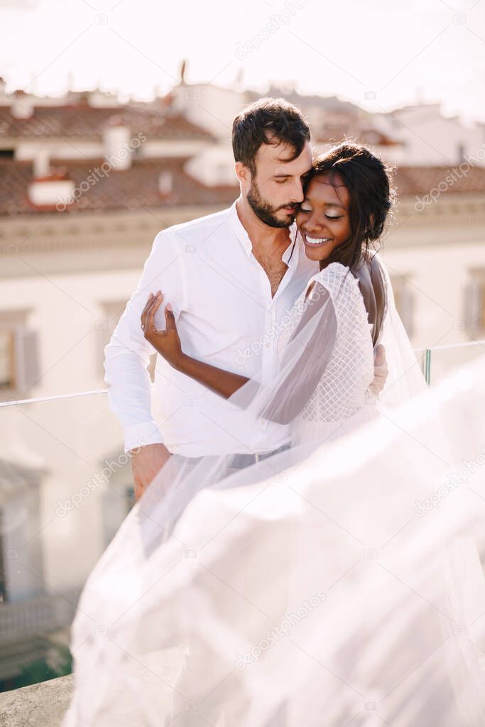 Destination fine-art wedding in Florence, Italy. Multiethnic wedding couple. African-American bride hugs Caucasian groom on the roof with cityscape view.