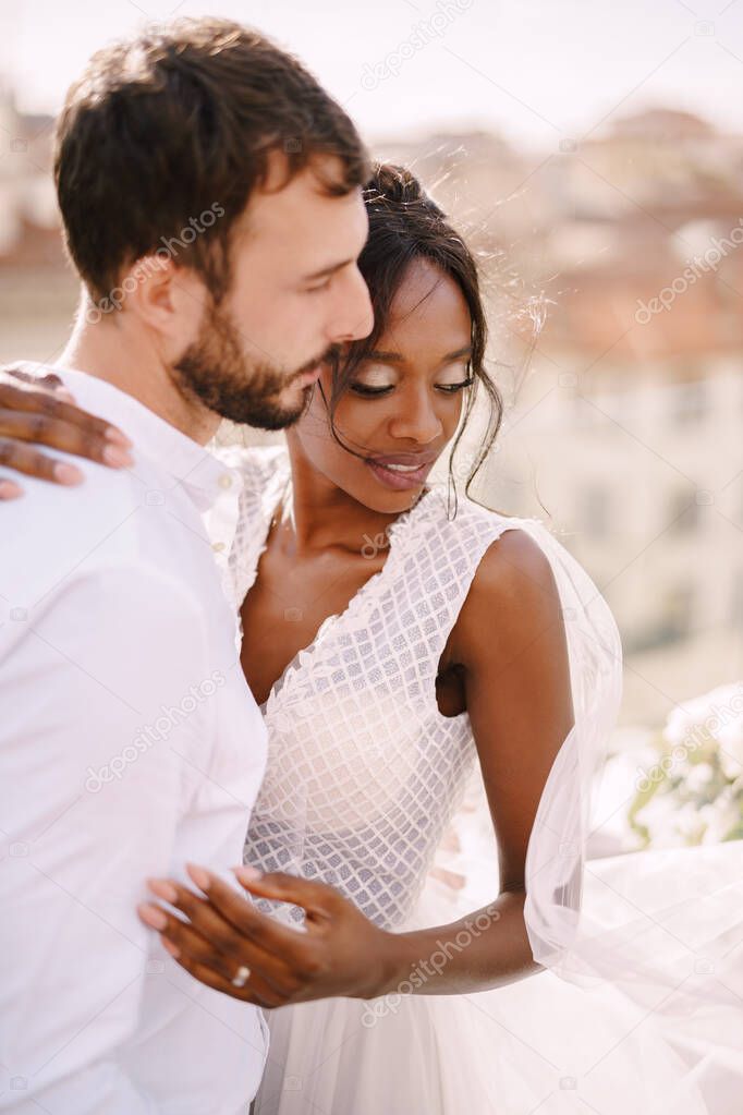 Destination fine-art wedding in Florence, Italy. Interracial wedding couple. Caucasian groom and African-American bride cuddling on a rooftop in sunset sunlight.