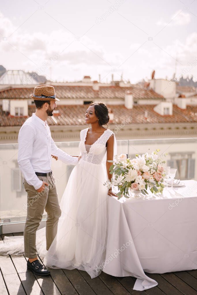 Interracial wedding couple. Destination fine-art wedding in Florence, Italy. African-American bride and Caucasian groom with a beard and straw hat, near the table for a wedding dinner.