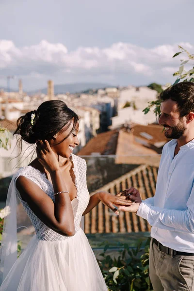 Multiethnic wedding couple - African-American bride and Caucasian groom. Groom puts a ring on brides finger. Destination fine-art wedding in Florence, Italy. Wedding ceremony on the roof