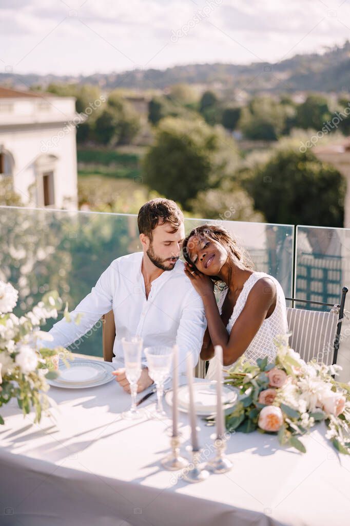 Destination fine-art wedding in Florence, Italy. African-American bride and Caucasian groom are sitting at the rooftop wedding dinner table overlooking the city. Multiracial wedding couple