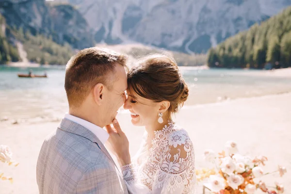 The newlyweds kiss on the spot for the ceremony, with an arch of autumn flower columns, against the backdrop of the Lago di Braies in Italy. Destination wedding in Europe, on Braies lake. — Stock Photo, Image