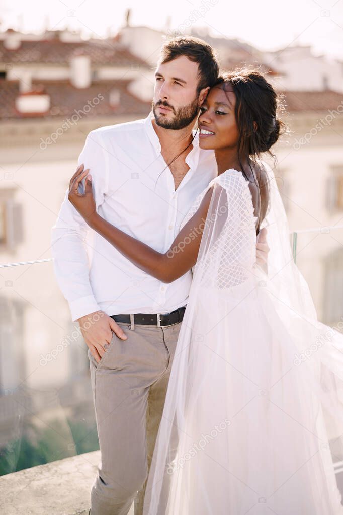 Interracial wedding couple. Destination fine-art wedding in Florence, Italy. African-American bride hugs Caucasian groom on the roof with cityscape view.