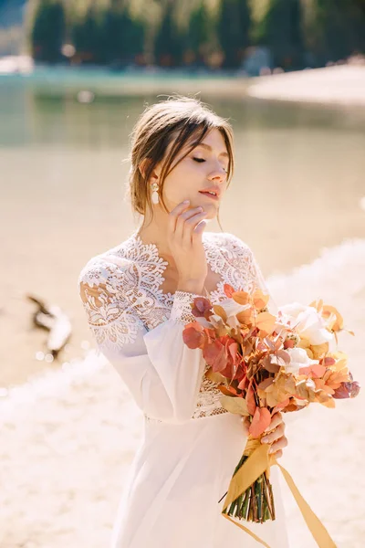 Όμορφη νύφη σε λευκό φόρεμα με μανίκια και δαντέλα, με ένα κίτρινο φθινοπωρινό μπουκέτο από αποξηραμένα λουλούδια και τριαντάφυλλα παιώνιας, στο Lago di Braies της Ιταλίας. Γάμος προορισμού στην Ευρώπη, στη λίμνη Braies. — Φωτογραφία Αρχείου