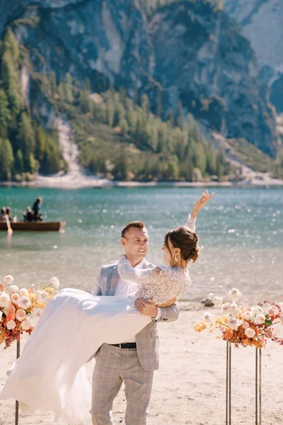 El novio rodea a la novia en brazos en el lugar de la ceremonia, con dos columnas de flores de otoño en lugar de un arco, con el telón de fondo del Lago di Braies en Italia. Boda en Europa, en el lago Braies . —  Fotos de Stock