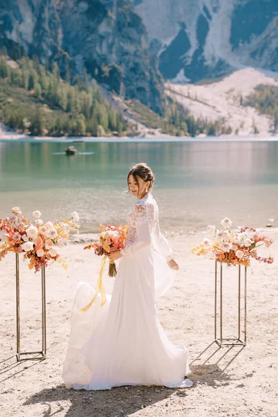Mooie bruid in een witte jurk met mouwen en kant, met een geel herfstboeket op de achtergrond van de boog voor de ceremonie, in Lago di Braies in Italië. Bestemmingsbruiloft in Europa, aan het Braies meer. — Stockfoto