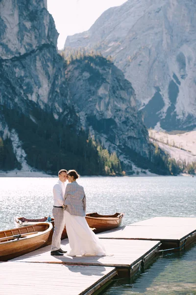 De bruid en bruidegom lopen langs een houten aanlegsteiger bij het Lago di Braies in Italië. Bruiloft in Europa, aan het Braies meer. Pasgetrouwden lopen, kussen, knuffelen op een achtergrond van rotsachtige bergen. — Stockfoto