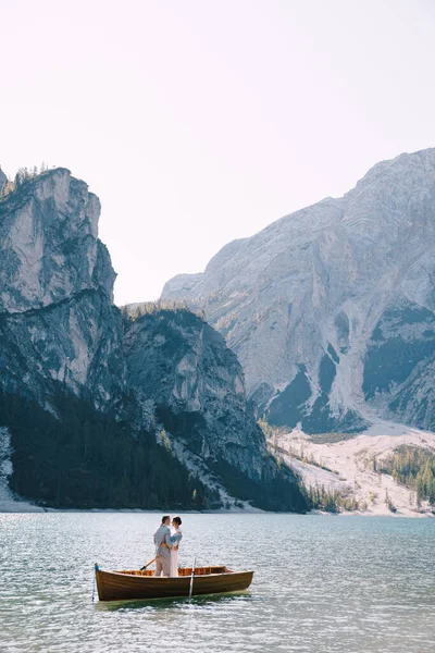 Νύφη και γαμπρός πλέουν με ξύλινη βάρκα, με κουπιά στη λίμνη Lago di Braies στην Ιταλία. Γάμος στην Ευρώπη - Οι νεόνυμφοι στέκονται αγκαλιασμένοι σε μια βάρκα. — Φωτογραφία Αρχείου