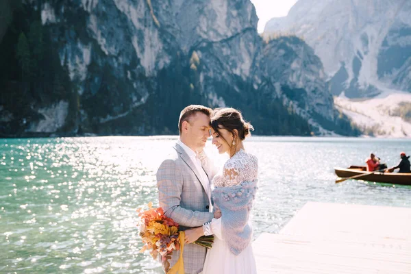 Das Brautpaar spaziert an einem hölzernen Bootsanleger am Pragser See in Italien entlang. Hochzeit in Europa, am Pragser See. Frischvermählte spazieren, küssen, umarmen sich vor dem Hintergrund felsiger Berge. — Stockfoto