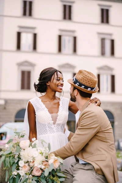 Multiracionális házaspár. Esküvő Firenzében, Olaszországban. Afrikai-amerikai menyasszony fehér ruhában, hosszú fátyollal és csokorral, és fehér vőlegény homokos dzsekiben és szalmakalapban.. — Stock Fotó