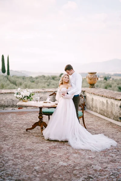 Esküvő egy régi borászati villában Toszkánában, Olaszországban. Egy házaspár áll az asztal mellett egy esküvői vacsorára, a vőlegény derekánál fogva öleli meg a menyasszonyt.. — Stock Fotó