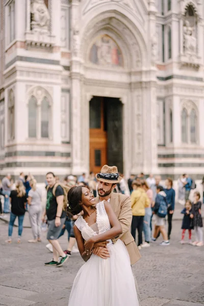 Pasangan pernikahan antar ras. Pernikahan di Florence, Italia. Pengantin pria kaukasia berpelukan dari belakang pengantin Afrika-Amerika di Piazza del Duomo. Stok Lukisan  