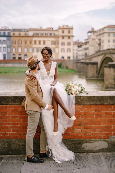 Fajok közötti házaspár. Esküvő Firenzében, Olaszországban. Egy afro-amerikai menyasszony ül a téglafalon, és a fehér vőlegény ölelgeti. Arno folyó töltése, kilátással a városra és a hidakra — Stock Fotó