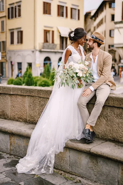 Interracial wedding couple. Wedding in Florence, Italy. African-American bride in a white dress with a long veil and a bouquet, and Caucasian groom in a sandy jacket and straw hat. — Stock Photo, Image