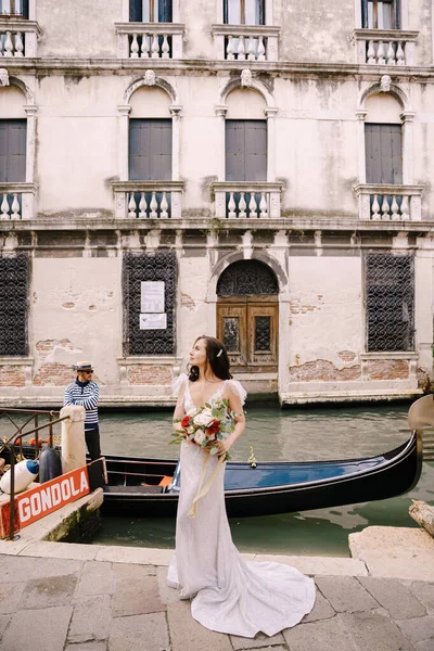 Venice, Italy - 04 october 2019: Venice wedding in Italy. 흰색 드레스를 입고 기차를 타고 손에 부케를 든 신부가 베네치아의 좁은 운하에 정박 해 있는 곤돌라 근처 부두에서 있다. — 스톡 사진