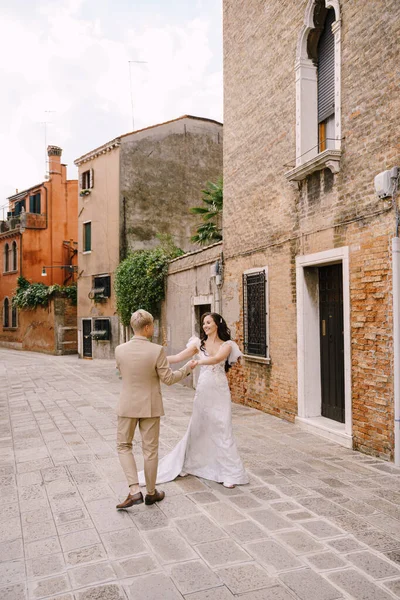 意大利在威尼斯的婚礼。新娘和新郎沿着城里荒废的街道散步.新婚夫妇拥抱着，跳舞，手牵着手，面对着美丽的红砖房子. — 图库照片
