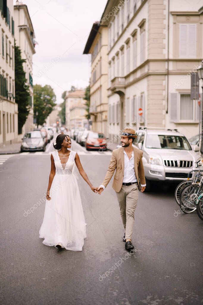 Multiethnic wedding couple. Wedding in Florence, Italy. African-American bride in a white dress and Caucasian groom in a hat are walking along the road among cars.