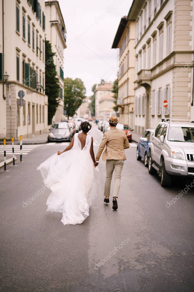 Wedding in Florence, Italy. Multiracial wedding couple. African-American bride in a white dress and Caucasian groom in a hat are walking along the road among cars.