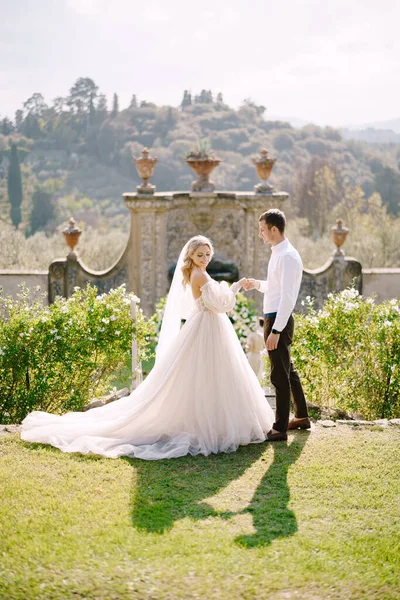 花嫁と新郎が公園を歩いている。イタリアのトスカーナにある古いワイナリーのヴィラで結婚式。白い花と緑で飾られた円形の結婚式のアーチ古代イタリアの建築の前で. — ストック写真