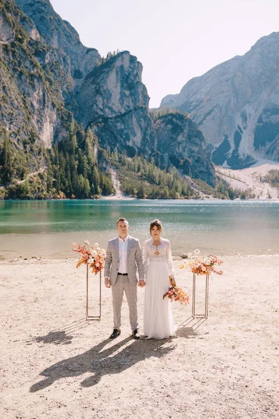 De bruid en bruidegom staan op hun plaats voor de ceremonie, met een boog van herfstbloemkolommen, tegen de achtergrond van het Lago di Braies in Italië. Bestemmingsbruiloft in Europa, aan het Braies meer. — Stockfoto