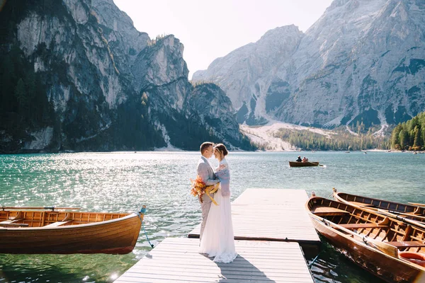 A menyasszony és a vőlegény sétálnak végig egy fa hajó dokk a Lago di Braies Olaszországban. Esküvő Európában, a Braies tónál. Friss házasok séta, csók, ölelés hátterében sziklás hegyek. — Stock Fotó