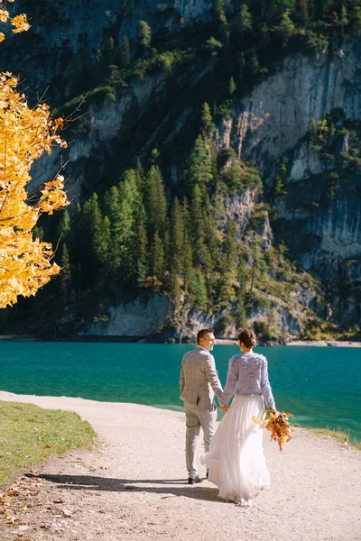 Bruid en bruidegom onder een herfstboom, met vurig geel blad, in het Lago di Braies in Italië. Bestemmingsbruiloft in Europa, aan het Braies meer. Geliefden die pas getrouwd zijn, lopen elkaar achterna.. — Stockfoto