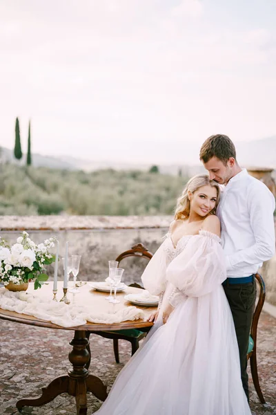 Casamento em uma antiga vinícola villa na Toscana, Itália. Um casal de casamento está de pé perto da mesa para um jantar de casamento, o noivo abraça a noiva pela cintura . — Fotografia de Stock